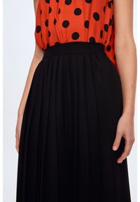 Gonna plissettata nera con vita alta. Il tessuto sembra liscio. Un top rosso con pois neri è parzialmente visibile sopra la gonna.