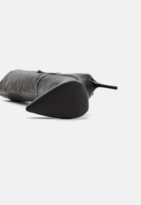 Svart stiletto-klack med en texturerad, plisserad läder överdel. Spetsig tå med en slät svart sula. Generell strömlinjeformad design.