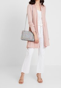 Ljusrosa lång kofta med strukturerad stickning, matchad med en vit topp och vida vita byxor. En flerfärgad vävd handväska och beige sandaletter fulländar looken.