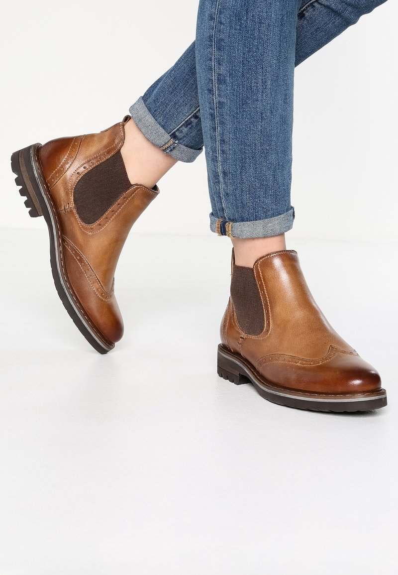 Bottes Chelsea en cuir marron avec détails brogue portées avec un jean bleu retroussé sur fond blanc.
