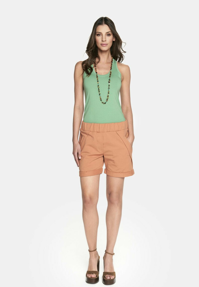 Zielony top na ramiączkach, pomarańczowe szorty z paskiem w talii, zawinięte mankiety, kieszenie boczne oraz brązowe sandały na platformie. Naszyjnik z ciemnymi koralikami.