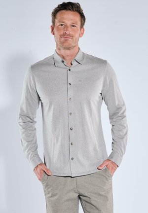 Mann trägt ein hellgraues, langärmeliges Hemd mit Knöpfen und eine beige Hose, steht mit den Händen in den Taschen vor einem einfarbigen Hintergrund.