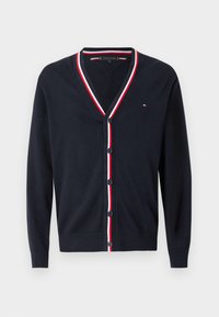 Ναυτικό πουλόβερ με V λαιμόκοψη, λευκές και κόκκινες ρίγες κατά μήκος του γιακα, έξι κουμπιά μπροστά και ένα μικρό λογότυπο στο στήθος.