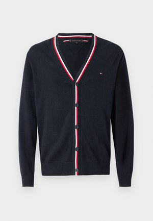 Ναυτικό πουλόβερ με V λαιμόκοψη, λευκές και κόκκινες ρίγες κατά μήκος του γιακα, έξι κουμπιά μπροστά και ένα μικρό λογότυπο στο στήθος.