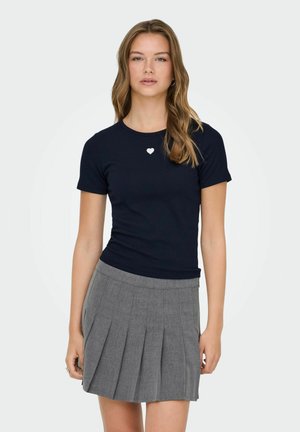 Junge Frau mit welligem blondem Haar, die ein eng anliegendes marineblaues T-Shirt mit kleinem weißem Herz und einen plissierten grauen Rock trägt.