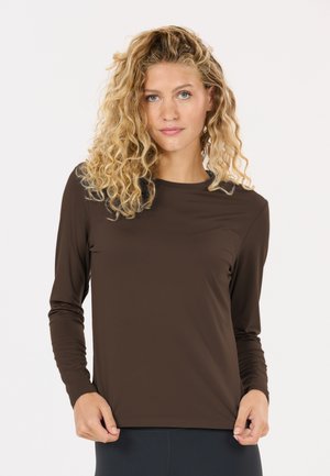Junge Frau mit lockigem blondem Haar, die ein braunes Langarmshirt trägt und den Saum hält, vor einem einfarbigen hellen Hintergrund.