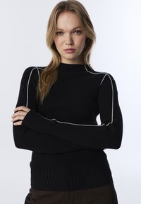 Jeune femme aux cheveux blonds portant un pull noir à côtes avec des bordures blanches, se tenant les bras croisés devant un fond uni.