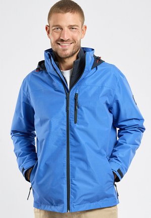 Homme souriant aux cheveux courts portant une veste bleue zippée les mains dans les poches, debout devant un fond clair uni.