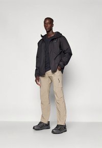 Černá voděodolná bunda s kapucí, doplněná světle béžovými cargo kalhotami s kapsami a zipy a tmavými turistickými botami.