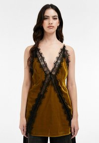 Femme portant un haut sans manches en velours moutarde avec un décolleté en V profond et une bordure en dentelle noire, associée à un pantalon sombre, se tenant devant un fond blanc uni.