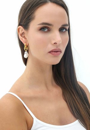 Orecchini in tono oro con un cerchio texturizzato e una perla bianca irregolare pendente. Il modello ha i capelli scuri e lisci e uno sfondo neutro.