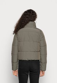 Veste rembourrée en vert olive, avec un col montant, un design matelassé et une coupe courte. Le matériau semble lisse et léger.