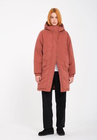 Padded coat w stonowanej terakocie z wysokim kołnierzem i kapturem. Wyposażony w zamek błyskawiczny, kieszenie z przodu i elastyczne mankiety. Nosi się go na czarnych spodniach.