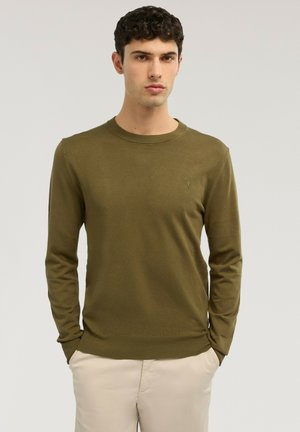 Giovane uomo con maglione a maniche lunghe verde oliva e pantaloni beige, in piedi con le mani parzialmente nelle tasche contro uno sfondo semplice.