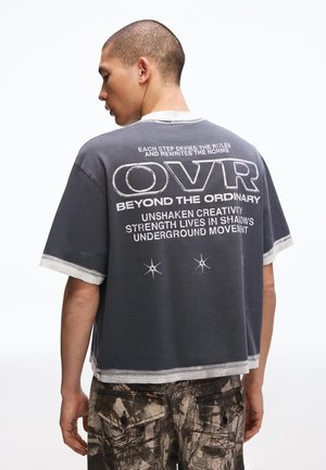 Junger Mann, von hinten gesehen, trägt ein dunkles, kurz geschnittenes T-Shirt mit weißem Text und Grafik, kombiniert mit Tarn-Cargohosen.