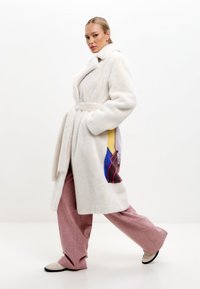 Vit fuskpälsjacka med bälte i midjan, med färgglada geometriska mönster på ena sidan, bärs över rosa vida byxor.