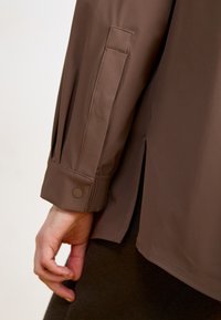 Giacca marrone leggera con design delle maniche plissettate, polsini con bottone e spacchi laterali. Texture liscia e stile minimalista.