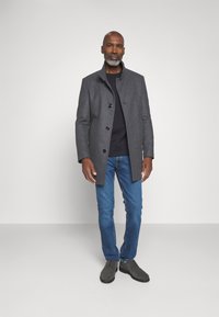 Cappotto di lana grigio con colletto alto, bottoni neri, abbinato a una maglia nera, jeans blu e scarpe grigio scuro, su uno sfondo neutro.