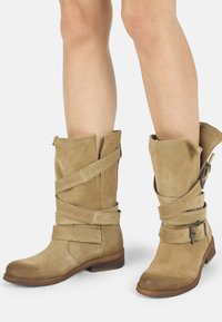Botas de ante color beige hasta la rodilla con puntera redondeada, que presentan tiras entrelazadas y un detalle de hebilla lateral sobre un tacón de madera apilada.