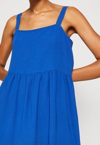 Vestido sin mangas azul con escote cuadrado, falda fruncida y una tela texturizada, que presenta una silueta relajada y fluida.
