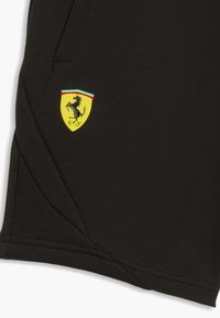 Tela negra con un logo de escudo Ferrari amarillo que presenta un caballo encabritado y los colores de la bandera italiana cerca del borde de un bolsillo cosido.