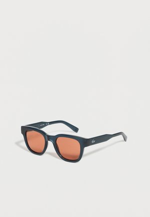 Lunettes de soleil rectangulaires noires avec verres teintés orange et logo Lacoste sur la branche, présentées sur fond blanc.