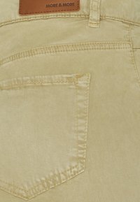 Détail de la poche arrière sur un pantalon beige avec couture visible et une étiquette en cuir marron portant la mention "MORE & MORE" au niveau de la taille.