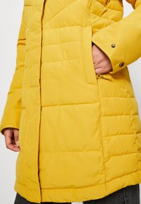 Närbild av en person som bär en vadderad gul jacka med en hand i sidofickan och tryckknappar på manschetten.