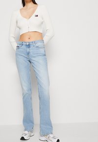 Donna che indossa un cardigan corto bianco con logo, jeans dritti blu chiaro e sneaker bianche, in piedi contro uno sfondo chiaro e uniforme.
