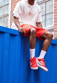 Punaiset lenkkarit, joissa on valkoiset raidat, valkoiset nilkkasukat, oranssit urheilushortsit ja valkoinen t-paita, jossa on graafinen kuvio, istumassa sinisellä kontilla.