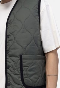 Gilet matelassé en vert foncé, avec des accents noirs, des boutons-pression et une poche avant. Texture lisse avec une surface à motifs.