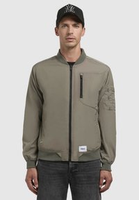 Khaki bomberjack met een rits aan de voorkant, ribboordkraag en -manchetten. Voorzien van een borstzak en een extra mouwzak met een logo-patch.