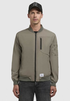 Khaki bomber dzseki elöl cipzárral, bordás gallérral és mandzsettával. Tartalmaz egy mellzsebet és egy további karzsebet logóval.