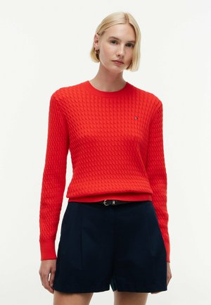 Vrouw met kort blond haar draagt een gestructureerde rode trui en zwarte korte broek tegen een effen lichte achtergrond.