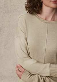 Maglione beige lavorato a maglia con scollatura rotonda, polsini a coste e dettagli delle cuciture sottili, che mette in mostra un tessuto morbido e testurizzato.