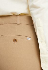 Béžové kalhoty vyrobené z texturované tkaniny, s jednou zadní kapsou na knoflík a malou bílou etiketou s nápisem "Polo". Viditelná světlá košile.