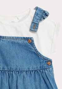 Das Denim-Kleid verfügt über einen weißen Baumwoll-T-Shirt-Aufsatz, einen blauen Denim-Stoff, geraffte Träger und zwei braune Knopfakzente an den Trägern.