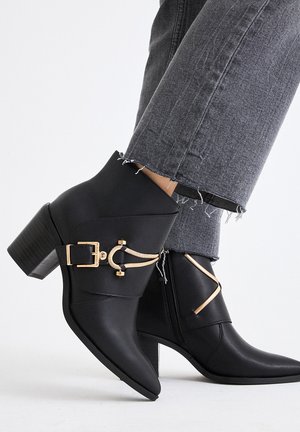 Botines negros con puntera puntiaguda, acentos de metal dorado y un tacón grueso, combinados con jeans gris desgastado.