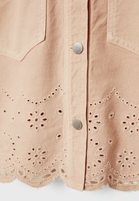 Tissu rose clair avec boutons-pression argentés, présentant des motifs floraux ajourés brodés et un ourlet festonné.