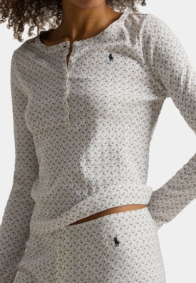 Vrouw draagt een bijpassend wit ribgebreid topje met lange mouwen en broek met kleine zwarte bloemenprint en donkerblauw paardenlogo op borst en heup.