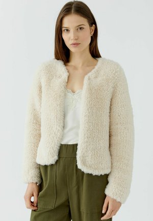 Jeune femme portant une veste duveteuse couleur crème, un haut blanc bordé de dentelle et un pantalon vert olive, debout devant un fond clair uni.