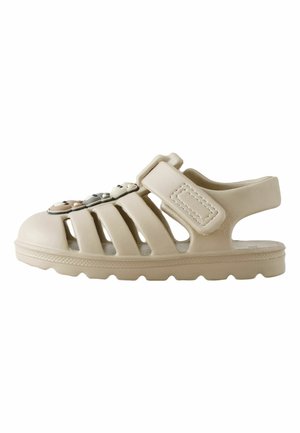 Beige sandal med lukket tå, justerbar rem og dekorative bjørneformede vedhæng på toppen, designet til afslappet brug og ventilation.