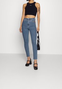 Jeans de mezclilla azul de tiro alto con un corte ajustado, que presentan un acento de cuentas coloridas en el bolsillo, combinados con un top corto negro y tacones gruesos.