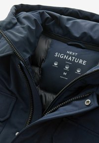 Navy blue down-filled jacket interior featuring a smooth texture, label with "NEXT SIGNATURE," and taped seams for waterproofing.