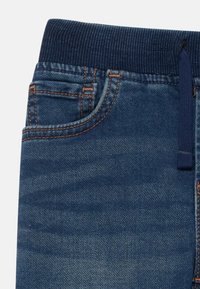 Pantalones vaqueros de mezclilla azul con cinturilla elástica acanalada y cordón de marino, mostrando un bolsillo frontal y costuras naranjas.