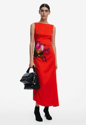 Rotes ärmelloses Kleid mit floralem Druck, figurbetonender Silhouette und seitlichen Rüschen. Kombiniert mit einer schwarzen Handtasche mit strukturierten Akzenten.