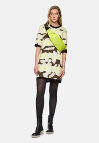 Übergroßes Kleid mit Camouflage-Muster in Beige, Braun und Neon-Gelb; kombiniert mit schwarzen Strumpfhosen und massiven schwarzen Plate Stiefeln. Neon-grüne Umhängetasche.