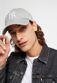 Gray baseball cap with white embroidered logo, curved brim, adjustable back strap. Worn by a person in a black denim jacket and white shirt.