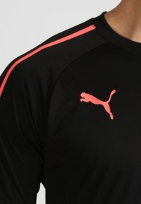 Camiseta atlética negra con acentos rosas a lo largo de los hombros. Presenta un prominente logo de Puma en color rosa en el pecho, fabricada con una tela suave y ligera.