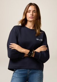 Donna con capelli castano chiaro lisci, che indossa una felpa blu scuro con la scritta "Sport" e jeans blu con dettagli in strass, con le braccia incrociate.